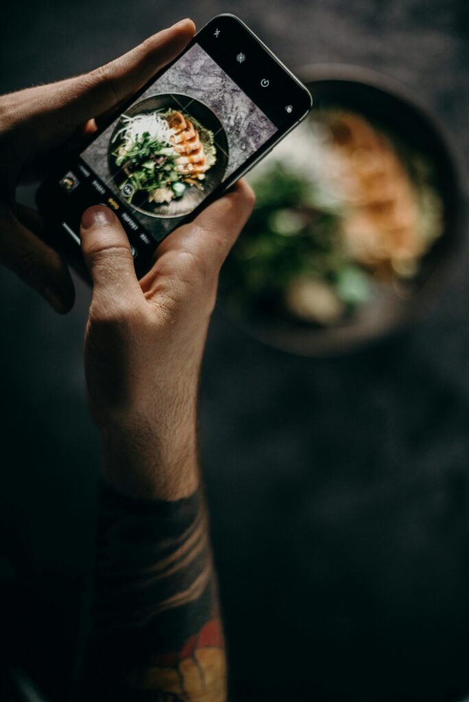 A person photographs a Japanese dish using a smartphone in a dimly lit setting.
